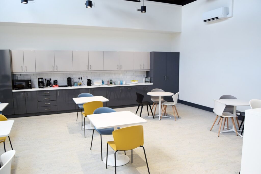 A modern canteen area with light gray cabinets, a tiled backsplash, and a variety of kitchen appliances. The room includes several round and square tables with colorful chairs, providing a comfortable dining space for employees.