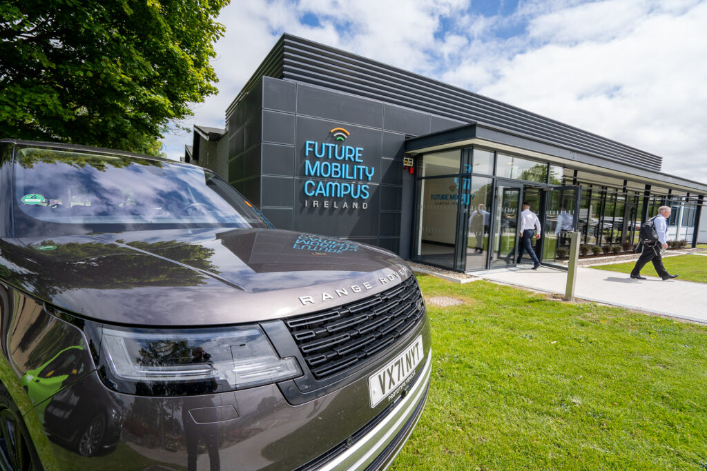 Future Mobility Campus Ireland (FMCI) office facility for smart mobility and autonomous vehicle research. Two men in suits enter and exit the modern, glass-fronted building with a Land Rover Defender test vehicle parked on the grass in front.