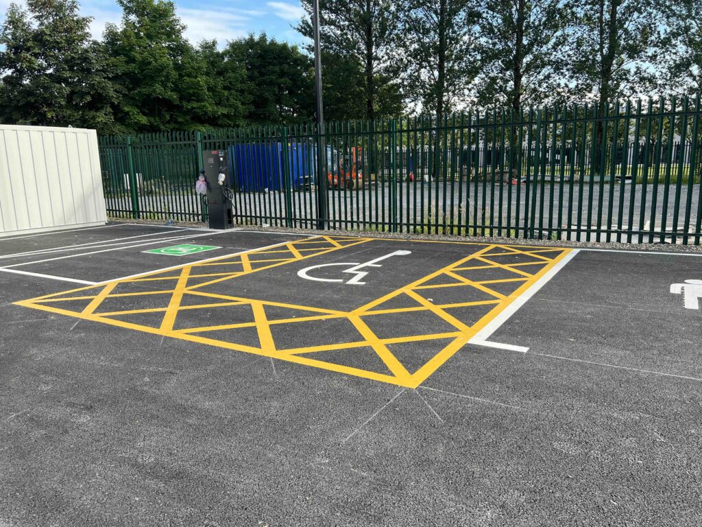 Designated accessible parking space and EV charging bay in the FMCI proving ground for connected and autonomous vehicles. Bright yellow road markings and an electric vehicle charging station are visible in the fenced testing area for sustainable transport and smart mobility solutions.