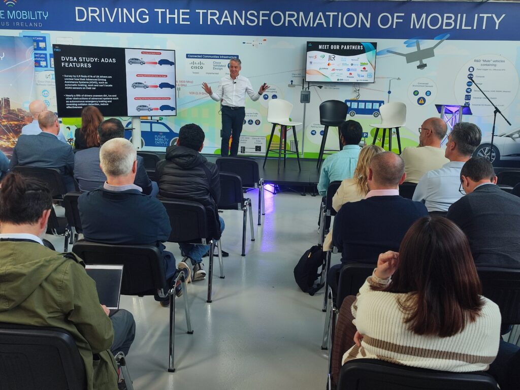 FMCI team member delivers opening address to an attentive audience in FMCI’s event venue, discussing a study on advanced driver-assistance systems (ADAS). Multiple screens behind him display data, images of connected vehicles, and partner logos.