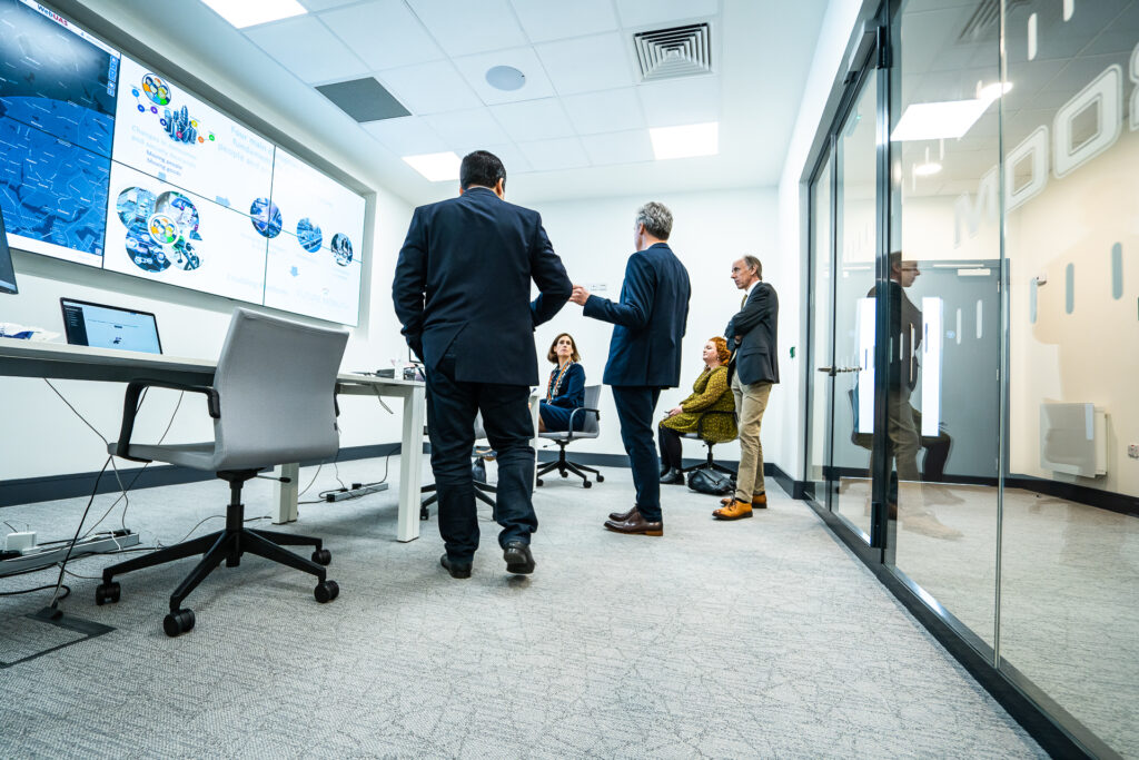 A group of professionals including three men and two women engage in discussion in FMCI's smart mobility control room. The control room features a large digital display wall of monitors showcasing mobility data visualisations, glass walls, a large table with laptops, and wired connections.