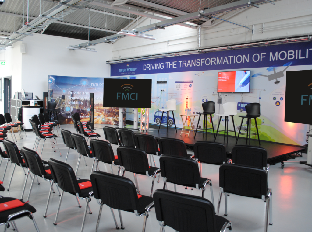 A modern event space in Shannon set up for a conference Future Mobility Campus Ireland, featuring a stage with high chairs, large digital screens, and rows of black chairs. The mural backdrop highlights smart mobility themes, including autonomous vehicles, drones, and connected transport technologies.