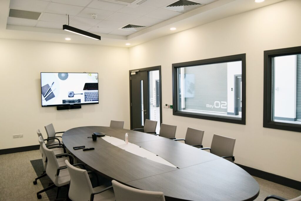 A professional conference room featuring a large oval table surrounded by ergonomic chairs. A wall-mounted screen displays a connected laptop interface, and the room is well-lit with contemporary lighting and large windows overlooking the workshop bays.