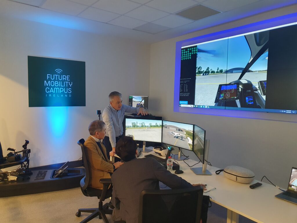 A smart mobility control center at FMCI, where professionals are monitoring advanced air mobility flight simulations on multiple large display screens. A presenter is explaining the data on-screen, which showcases a digital interface for testing autonomous aerial and ground mobility solutions. The immersive setup includes workstations, high-resolution monitors, and advanced analytics tools.