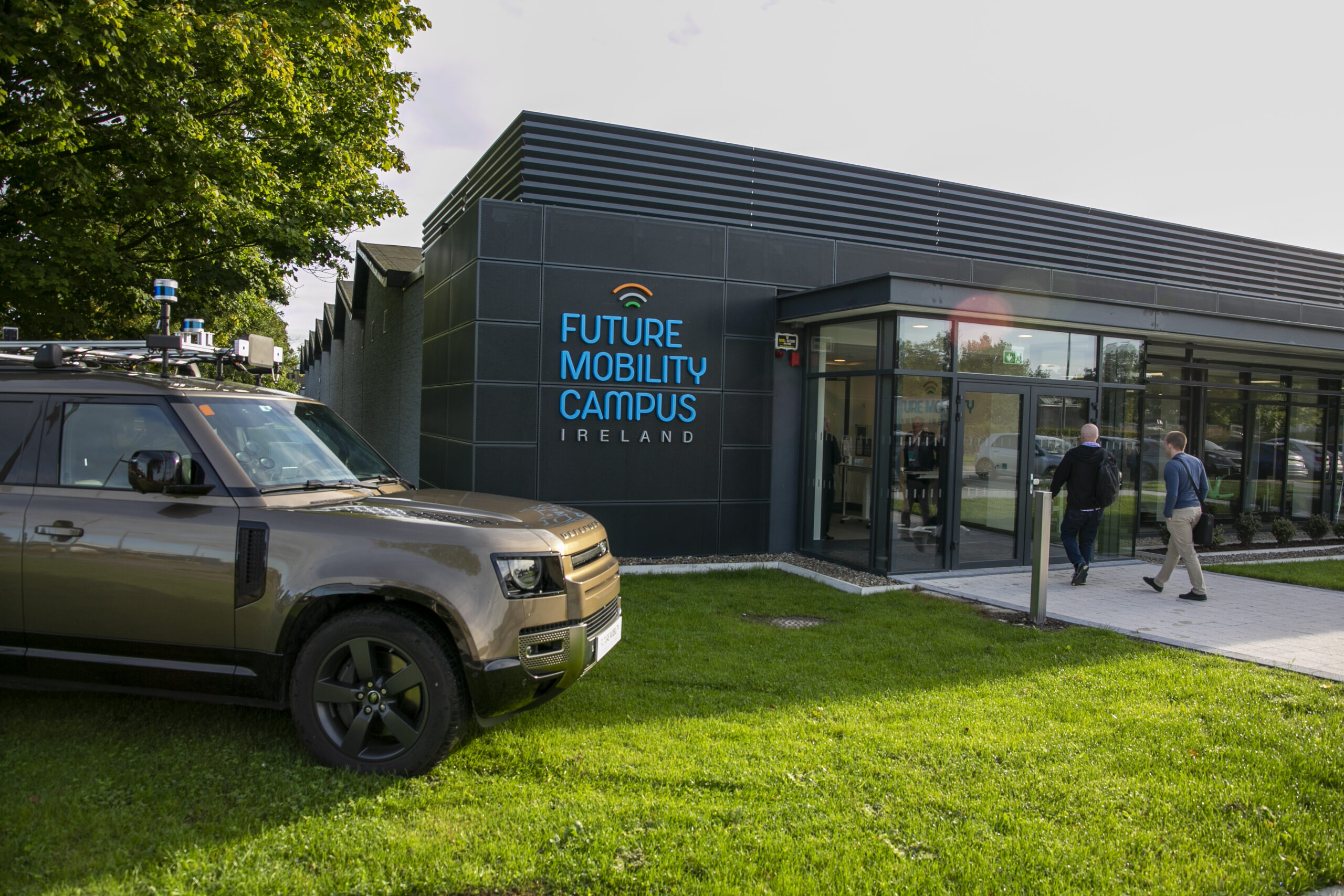 A modern black building with a sign reading 'Future Mobility Campus Ireland.' A Land Rover Defender, equipped with autonomous vehicle technology including sensors and cameras, is parked on the grass in front of the building.