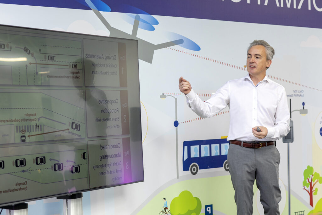 Speaker presenting at Future Mobility Campus Ireland with a clicker in hand, headset microphone, and large screen displaying smart mobility and autonomous driving concepts. The background features a mural illustrating urban mobility solutions.