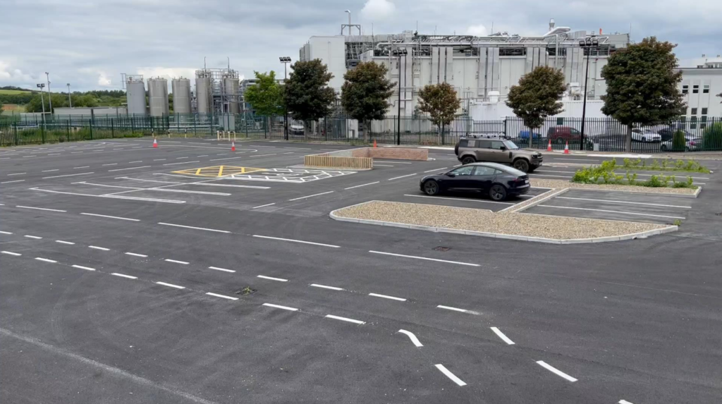 Test site and proving ground for connected and autonomous vehicles at Future Mobility Campus Ireland. A marked parking area with dedicated spaces and different conditions like gravel, vegetation, and smooth or cracked pavement are visible. A Tesla and Land Rover Defender are parked in the lot.