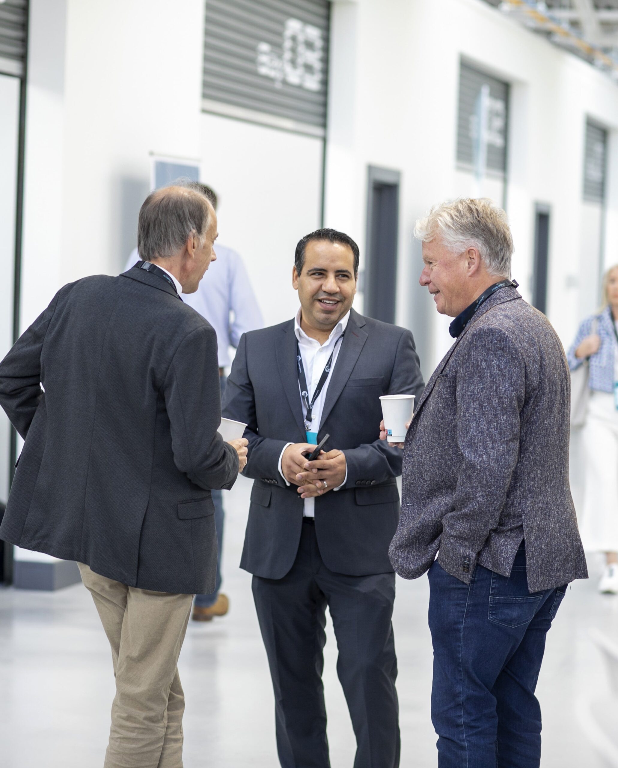 Three professionals networking at FMCI’s state-of-the-art facility, each holding a cup of coffee. Behind them, bright white walls, high ceilings, and labeled workshop bays create a sleek, industrial environment.