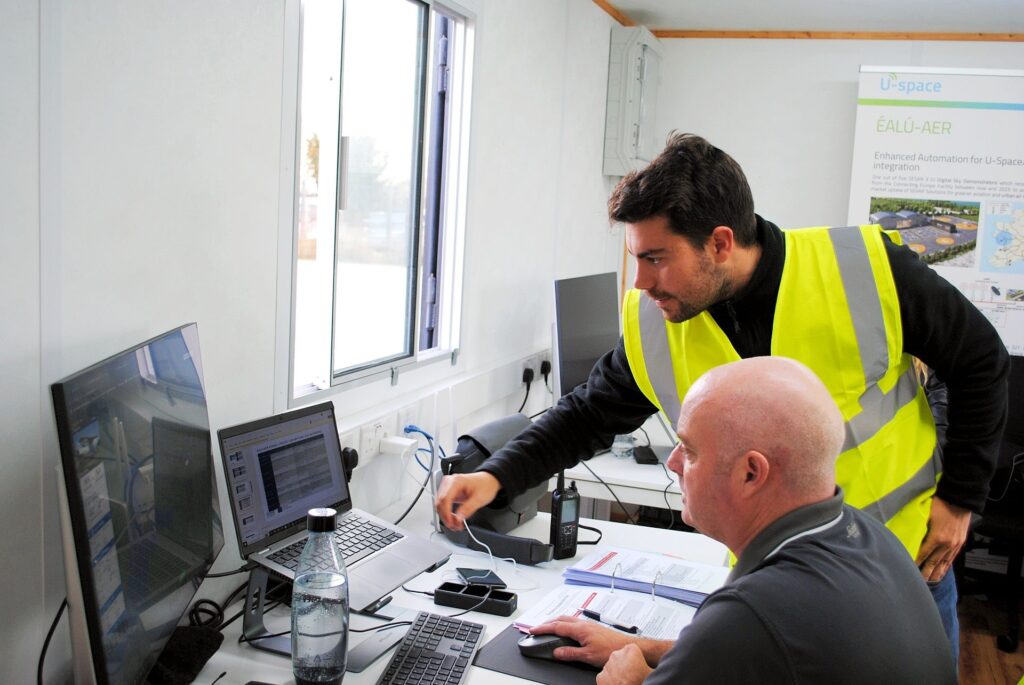 Two professionals, one in a high-visibility vest, work together inside the FMCI Drone Port control centre, monitoring flight operations for drone testing. One man sits at a desk, typing on a laptop while reviewing printed documents, while the other leans over, pointing at the screen. Multiple monitors display data, and a sign in the background references U-space and enhanced automation for drone integration, indicating an advanced air mobility testing environment.