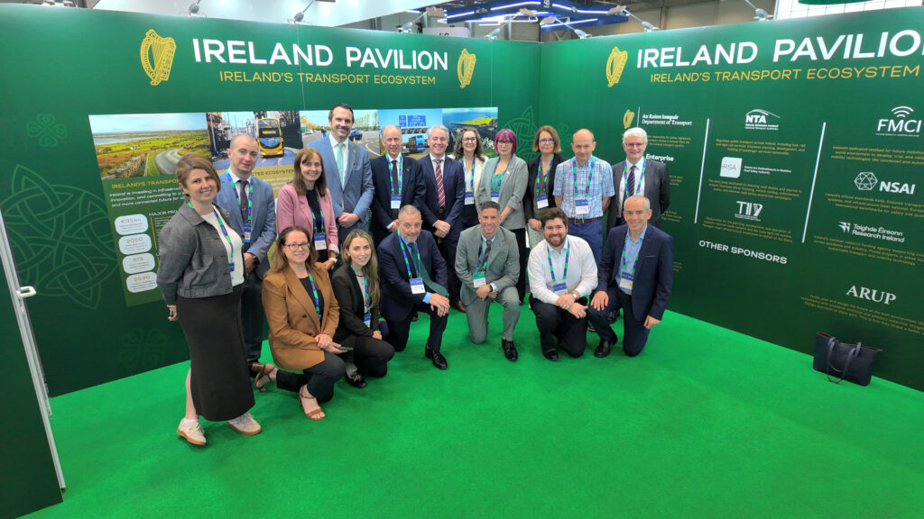A group of professionals pose in two rows for a photo. They are in the Ireland Pavilion stand at the TRA 2026 conference. A row in the back is standing while the front row kneels on a green carpet. Behind them, a dark green conference stand can be seen with the words "Ireland Pavilion: Ireland's transport ecosystem."